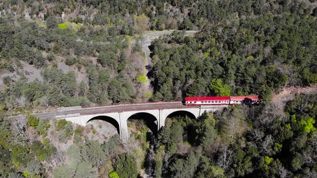 Aerial: red and white old train in a forest crossing an ancient viaduct in southern France