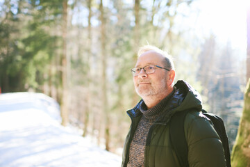 Outdoor portrait of middle age 55 - 60 year old man hiking in winter forest, wearing warm jacket and black backpack - 543449926