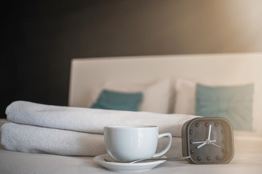 Soft light and Blurred image,A black alarm clock sits next to a white ceramic coffee cup on the bed in the morning as a cup with coffee prepared for guests to use after waking up in the morning.