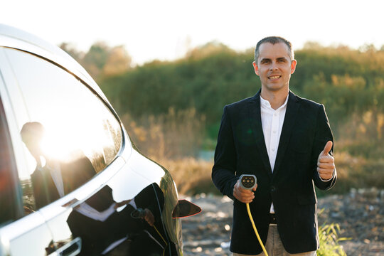 Portrait of caucasian businessman standing near electric charging station looking at camera and showing thumb up. Male holds a charging cable type 2 mennekes in his hands. Eco-friendly transport