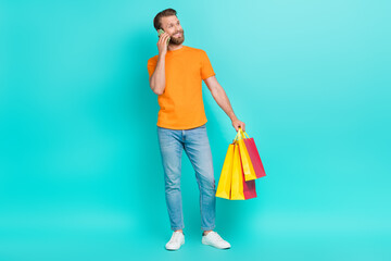 Full length photo of positive guy with brown hair wear orange t-shirt talking phone look empty space isolated on teal color background
