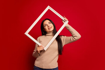 Pretty woman holding frame. Surprise. Winning success happy girl celebrating being a winner. Image of caucasian female model on red studio background. Human facial emotions concept.