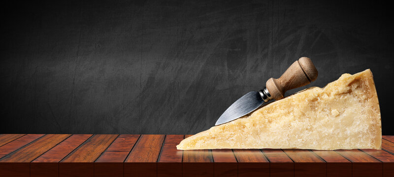 Close-up of a slice of Italian Parmesan cheese and old knife on a wooden table top for products display and a blank chalkboard on background.