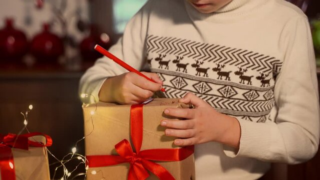 Close up child hand paints gift box New year present packaging. Making surprise for family. Kid dreaming miracle. Christmas eve holiday fairy lights and ringing bells.