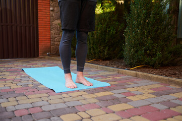 Cropped view of an active man, sportsman, a yogi standing barefoot on a blue mat while practicing yoga outdoors at sunset. Copy advertising space. Active healthy lifestyle. Fitness. Sport. Workout