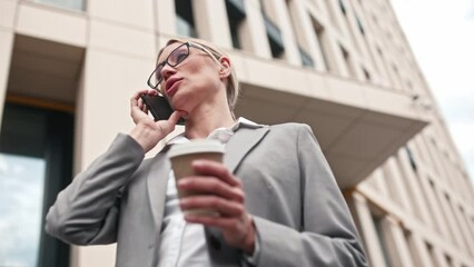 Successful businesswoman talking on phone holding cup of coffee wearing smart clothes and glasses. Attractive blonde concentrated female working staying outside. Businesswoman concept.