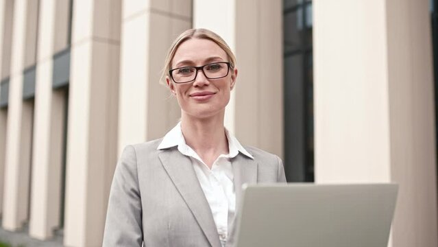 Attactive successful kind businesswoman staying outside wearing suit and glasses with laptop. Stunning pretty wealthy lady looking at camera smiling. Businesswoman close-up concept.