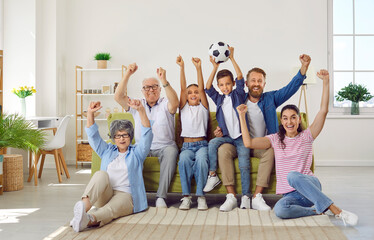 Modern active family of three generations is watching football match on TV at home together. Elderly and young family members sit on sofa and excitedly raise their hands in joy of scoring goal.