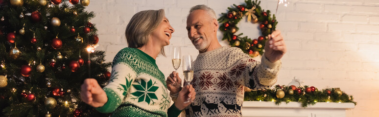 happy mature couple in sweaters holding glasses of champagne and sparklers on christmas eve, banner