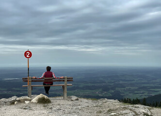 Wanderer (weiblich) mit roter Jacke sitzt auf einer Bank mit einem Schild mit der Nummer 2 und...