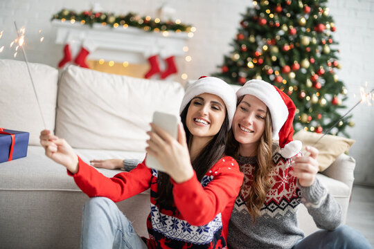 Cheerful same sex couple in santa hats taking selfie on smartphone and holding sparklers at home