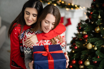 Positive woman hugging girlfriend with present near christmas tree at home