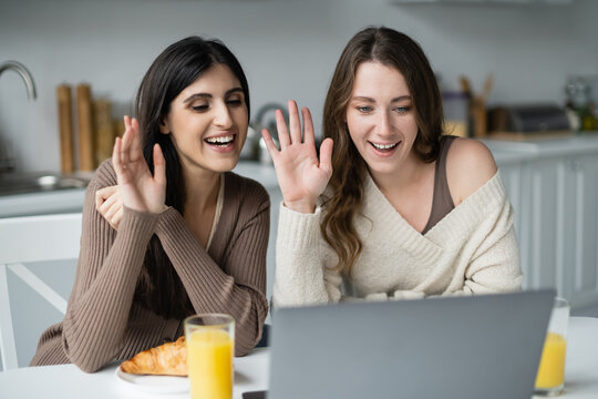 Positive lesbian couple having video call on laptop near breakfast in kitchen - Powered by Adobe