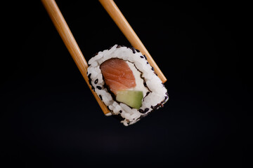 salmon cheese and avocado uramaki roll held by chopsticks on a black background