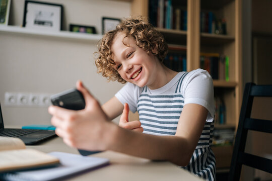 Cheerful Curly Little Girl Holding And Using Mobile Phone Showing Thumb Up, Browsing Social Network Apps, Surfing Internet And Scrolling News Feed, Reading Electronic E-book At Table With Laptop.