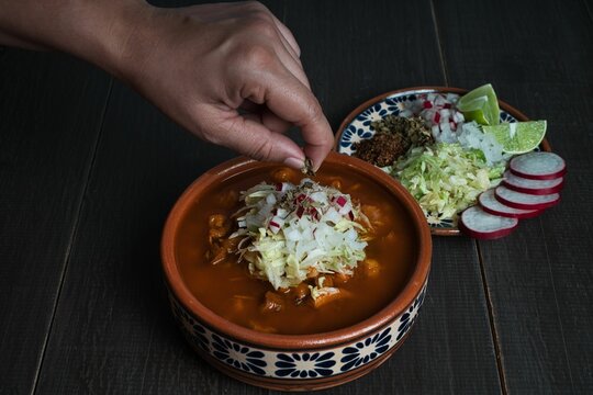 Closeup Shot Of A Hand Sprinkling Herbs On Pozole Rojo (Mexican Pork And Hominy Stew) On A Table