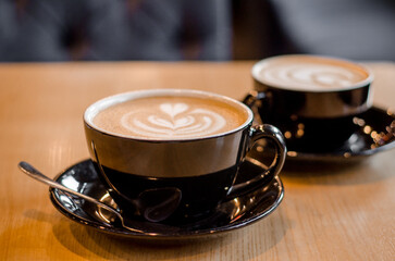 Two big black mugs with saucer containing latte or cappuccino coffee drink on a wooden table in cafe, lateart heart