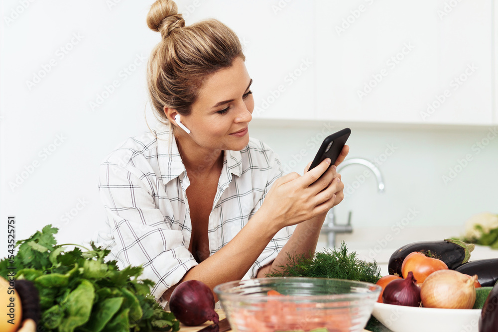 Wall mural Woman with wireless earbuds is using smartphone during cooking in modern white kitchen