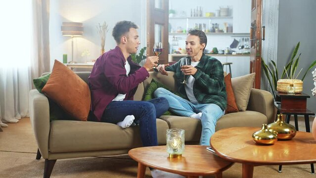Adorable male gay couple celebrating, making a toast and drinking wine together while sitting on a couch in a cozy stylish living room. Loving boyfriends having a date. LGBT relationship concept. 