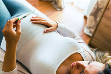 Fototapeta premium Pregnant temperature fever check. Pregnancy woman holding thermometer, check fever flu temperature. Pregnant girl sick. Concept of pregnancy, maternity, expectation for baby birth.