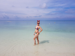 beautiful sexy fit woman in bikini and sunglasses standing in blue water on beach. Female wearing christmas reindeer headband. Tropical celebration of festive holiday and new year. Travel and vacation