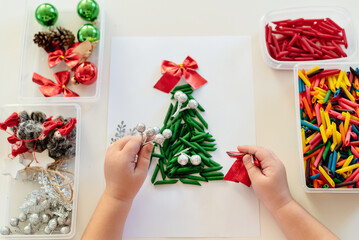 Children's hands creating Christmas tree from dyed pasta for the holiday of Christmas. Sensory play for childs. Holiday Art Activity for Kids, material Montessori, fine motor skills, sensory bin - obrazy, fototapety, plakaty
