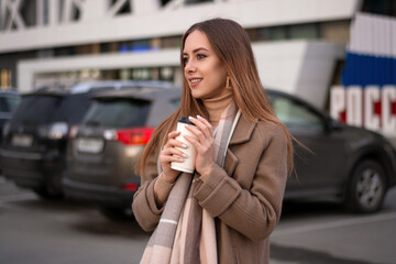 Fototapeta premium a beautiful girl in a coat holds a glass of coffee in her hands and walks around the city