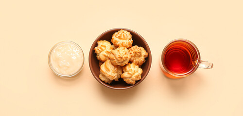 Bowl of tasty profiteroles, cream and cup of tea on beige background, top view