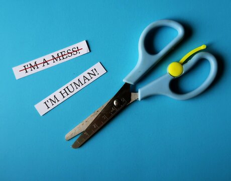 Scissors on blue background with handwritten two paper notes I AM A MESS, changed to I AM HUMAN - to overcome negative self talk or inner voice critic, change to positive thought to boost self esteem
