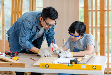 Asian professional male carpenter woodworker engineer dad in jeans outfit with safety gloves and goggles helping teaching young boy son using measuring ruler pencil marking stick in construction site