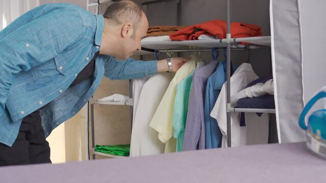 The man is choosing clothes from the closet.
Man with his clothes hanging in the closet is choosing clothes.
