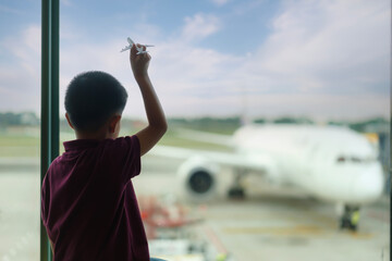 Asian Boy with airplane model at empty airport terminal waiting for departure looking out the window. Child in T-shirt and shorts stands at lounge waiting for plane flight. Family trip concept