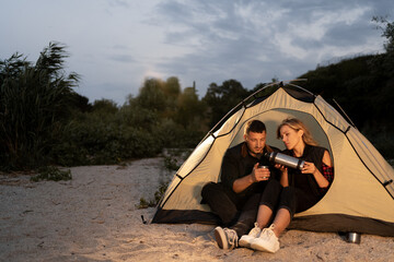 Night camping on the beach. Man and woman tourists are resting in front of a tent under the evening...