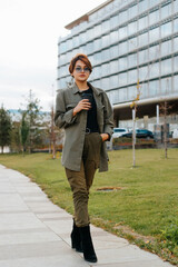 Slim stylish young Asian woman walking against backdrop of a glass building down street holding a cup of coffee and looking at camera, outdoors