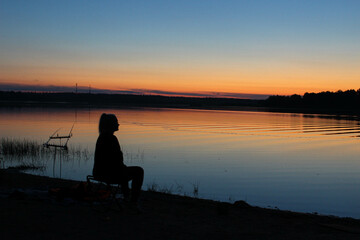 The girl sits thoughtfully on the evening bank of the river after sunset