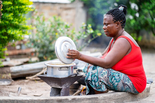 image of african mother cooking outside- black mother seated in front of a sauce pan with an opened lid