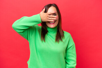 Middle-aged caucasian woman isolated on red background covering eyes by hands and smiling