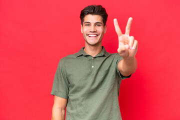 Young caucasian handsome man isolated on red background smiling and showing victory sign