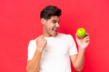 Young caucasian man with an apple isolated on red background celebrating a victory