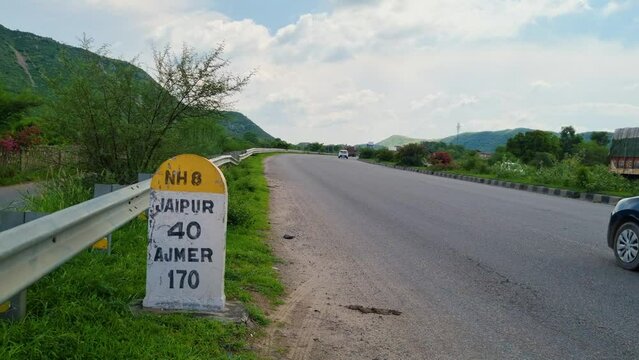Low shot showing the milestone on national highway NH8 showing the distance to Jaipur and Ajmer as cars zoom by on this highway surrounded by mountains