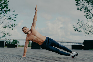Healthy man stands in abs side plank and raises arm, does exercises outdoor, has muscular body and...