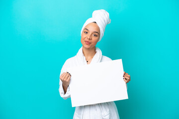 Young woman in a bathrobe with towel isolated on blue background holding an empty placard with happy expression