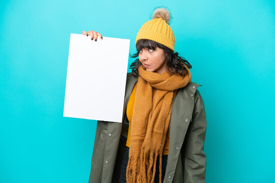 Young latin woman wearing winter jacket isolated on blue background holding an empty placard and looking it