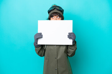 Skier latin woman with snowboarding glasses isolated on blue background holding an empty placard and hiding behind it