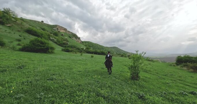 FPV sports drone around shot traditional culture Caucasian horseman with gun travers riding horse at summer mountain valley landscape. Aerial view male equestrian highlander at dramatic sky rocky 4k