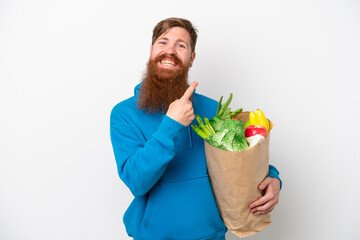 Redhead man with beard holding a grocery shopping bag isolated on white background pointing to the side to present a product