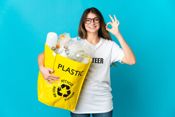 Woman holding a bag full of bottles to recycle isolated on blue background showing ok sign with fingers