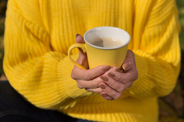 In female hands a yellow cup with cappuccino or cocoa, against the background of a yellow sweater, autumn concept