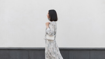 Beautiful elegant fashion girl with hairstyle in a fashionable flowers dress with a stylish bag walks near a white wall in the city, side view - obrazy, fototapety, plakaty