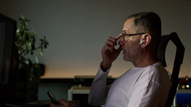 Middle Age Man Working From Home On Computer, Talking On Phone Using Wireless Earphones.